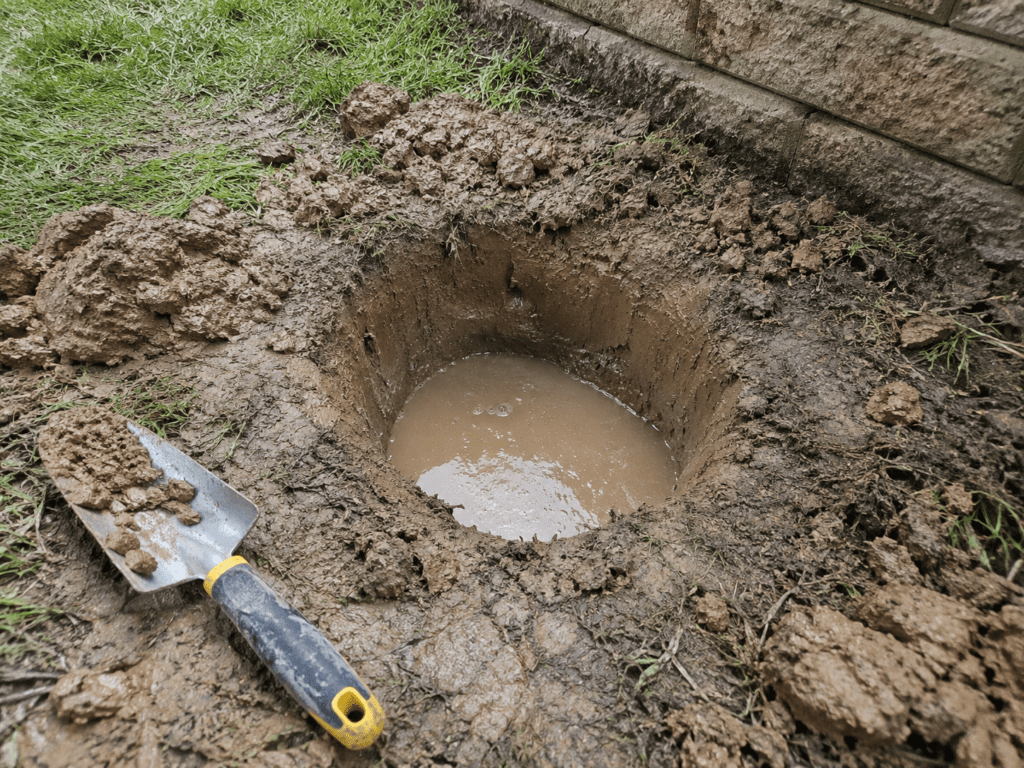 Small test hole with standing water in wet clayey soil near a retaining wall, showing poor site drainage after rain